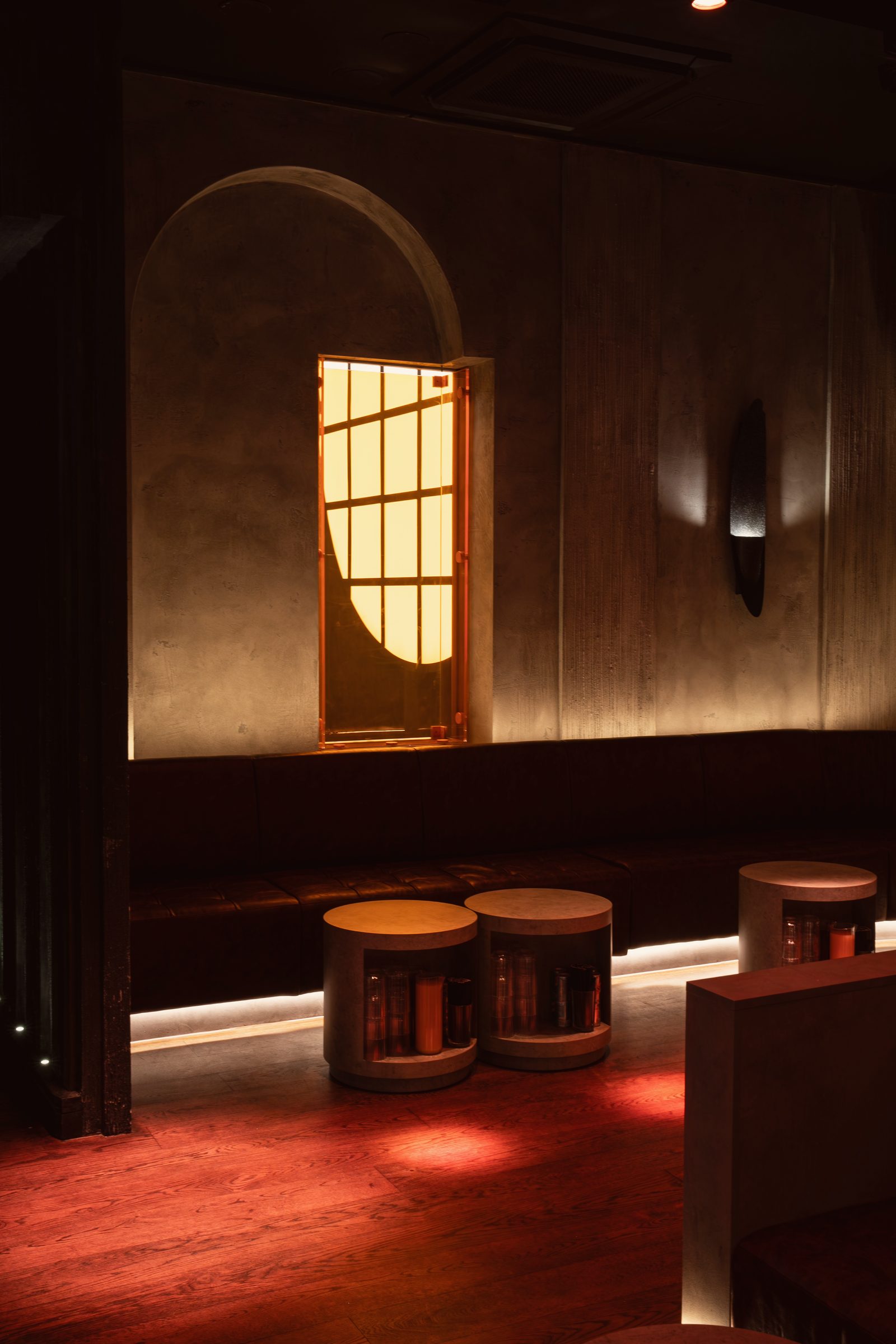An arched niche with a glowing amber window above a banquette and concrete drum tables